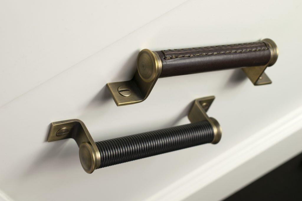 Two Bracket cabinet handles, one standing and one lay down. The bracket cabinet handle has a chunkier grip and is set on two L shapes legs with decorative domed collars. Shown in Chocolate leather with Fine Antique Brass.