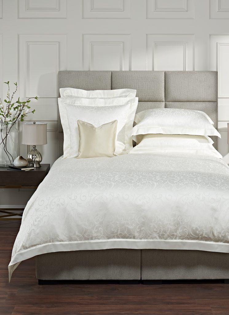 Luxury white linen bedroom with grey upholstered head board. The duvet has jacquard floral shapes and swirls. A very high end styled room with fresh delicate features.