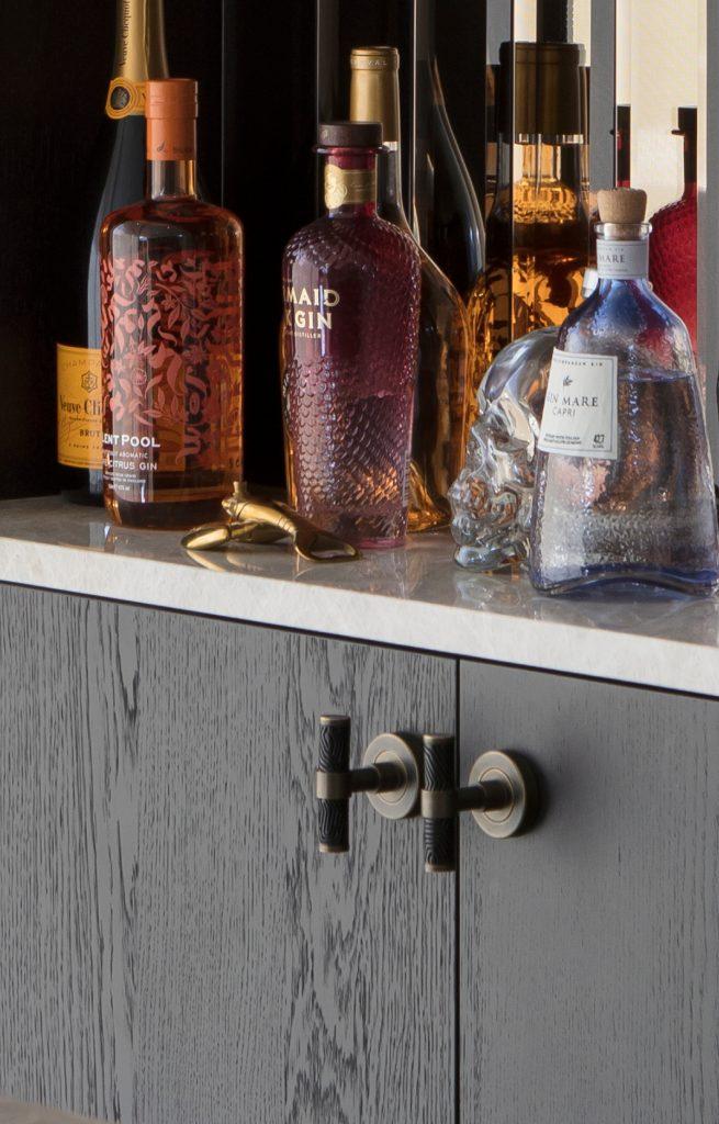 Luxury T Bar furniture handles by Turnstyle Designs have been used on dark work cupboard of this home bar. The T bars have The bar has a white quartz marble counter. Coloured spirit bottles decorate the counter top with cocktail making equipment.