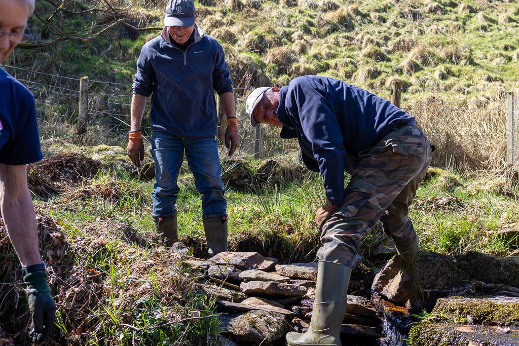 A group of older men help rebuild a wall that has collapsed in a stream, to direct the flow of water. This is at Ashcombe Gardens in Exmoor National Park.
