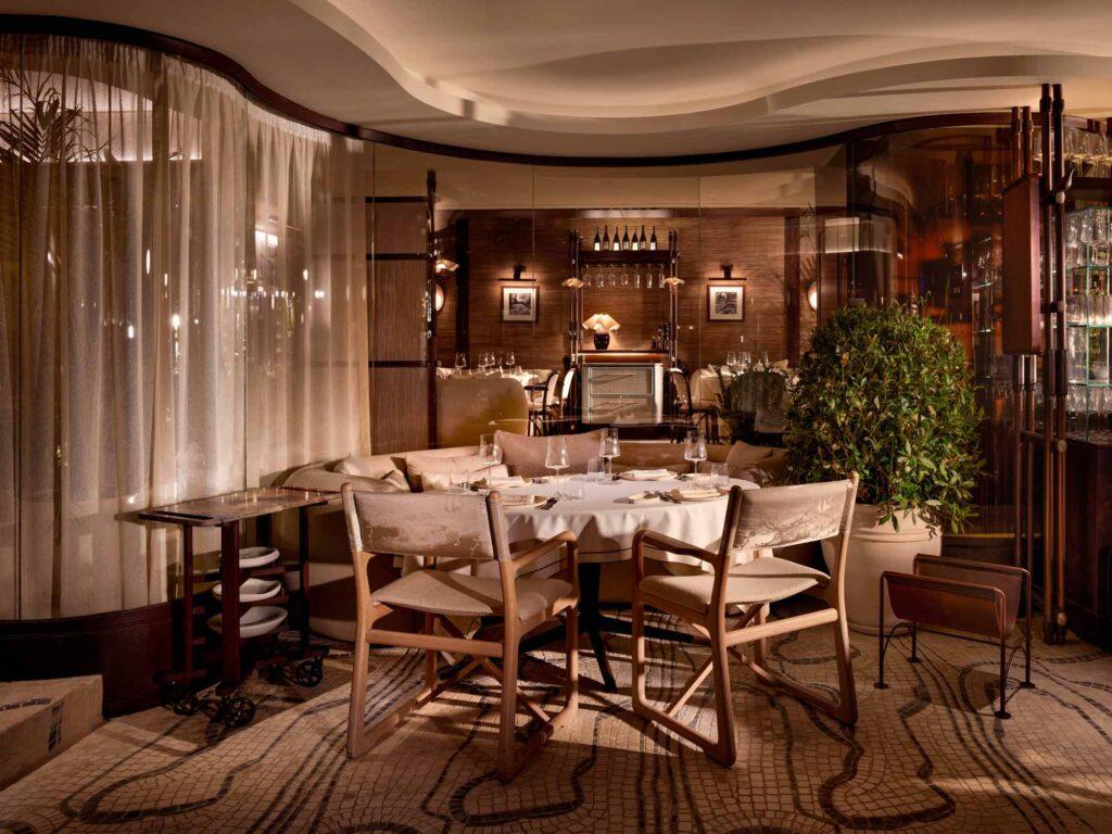 Elegant, softly lit restaurant interior with a round table set for fine dining. The space features neutral tones, natural wood chairs with canvas backs, plush upholstered seating, and curved glass walls with sheer curtains. A mosaic floor and potted greenery add texture and warmth, while shelves of glassware and wine bottles enhance the luxurious ambiance. The overall design evokes refined comfort and understated sophistication.